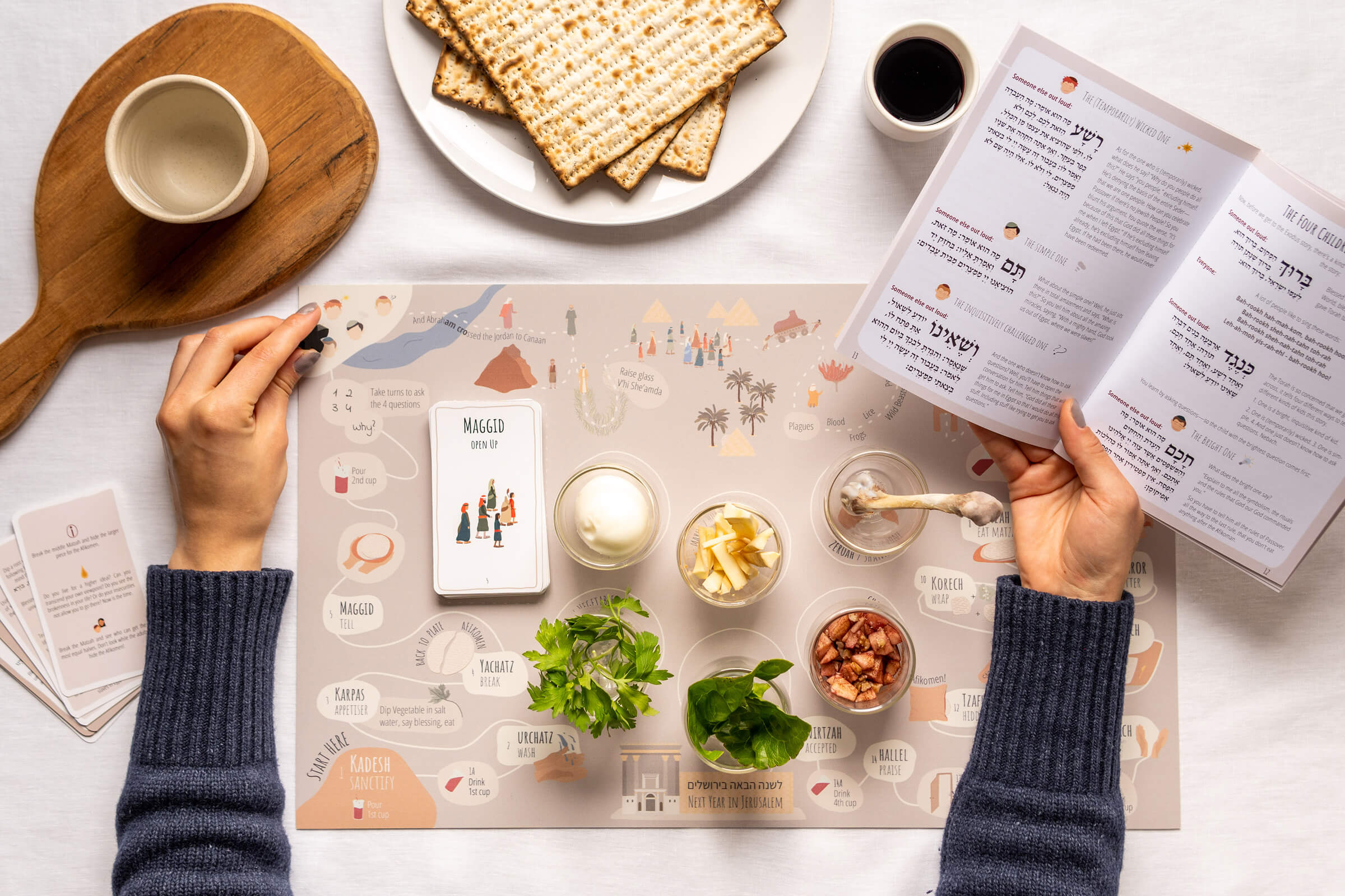Hands holding the Seder Companion book open beside the Seder board, matzah and wine