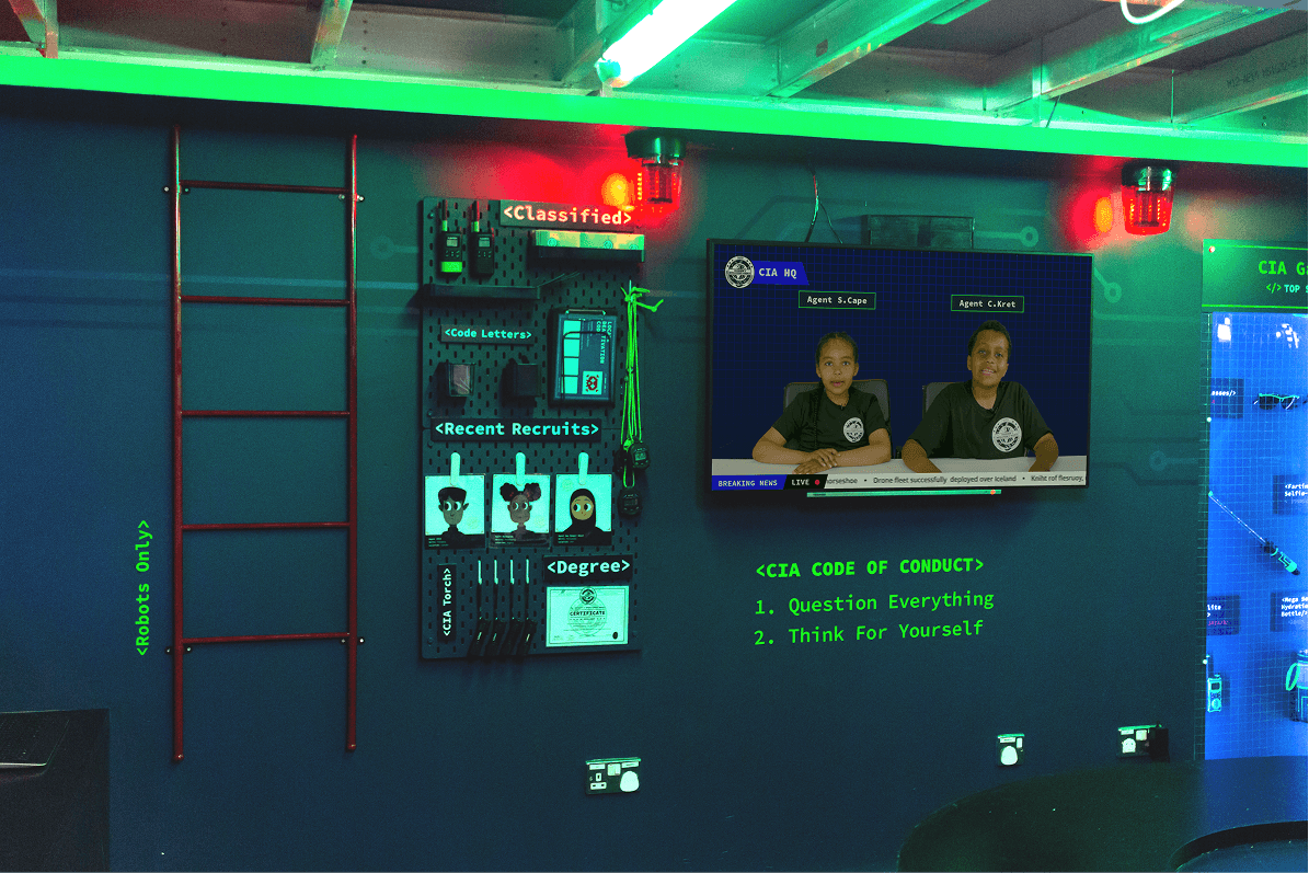 A wall-mounted screen showing a live CIA HQ broadcast, next to a code letters and recent recruits board