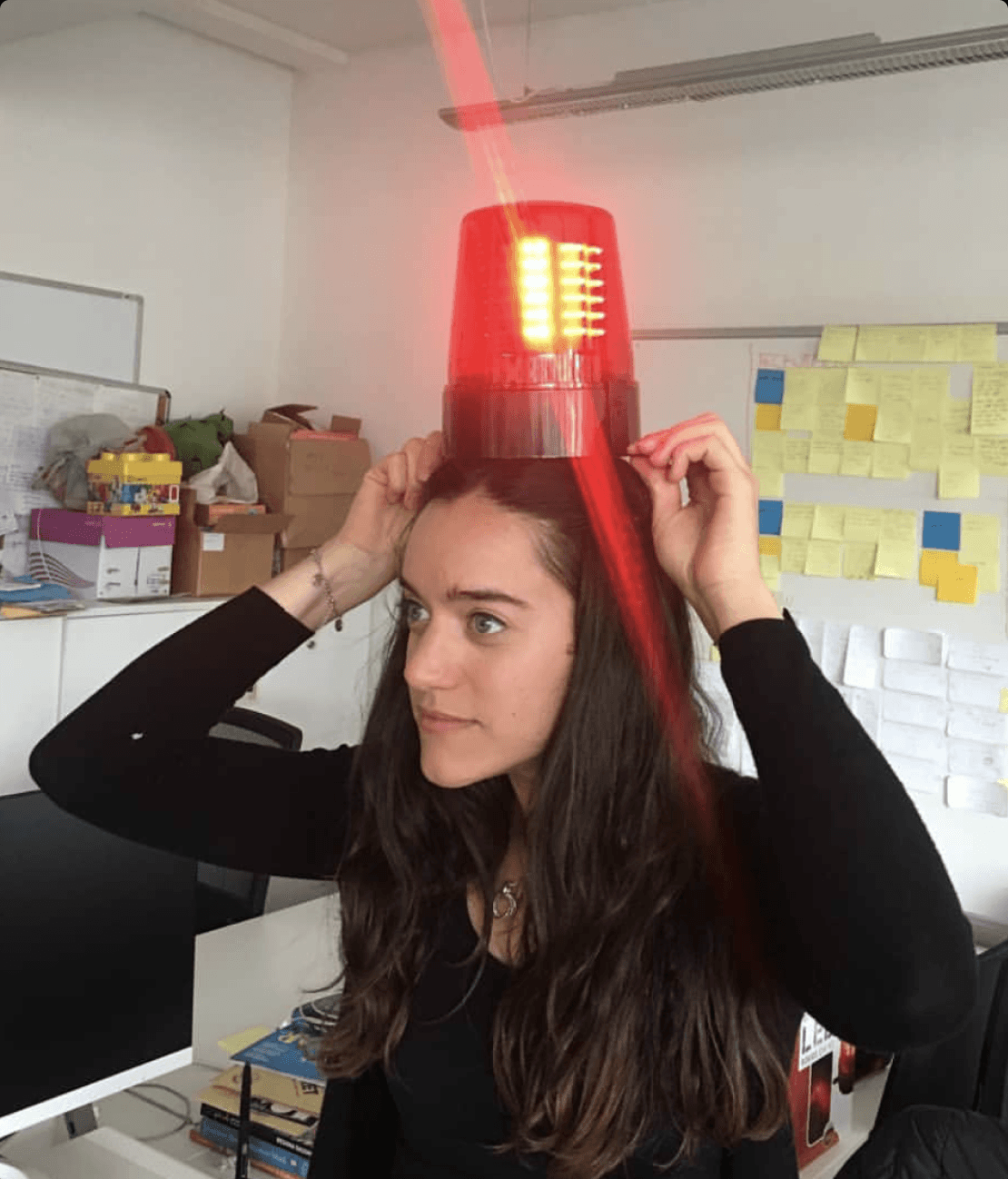 Lucrecia balancing a flashing red siren light on her head at her desk, surrounded by sticky notes