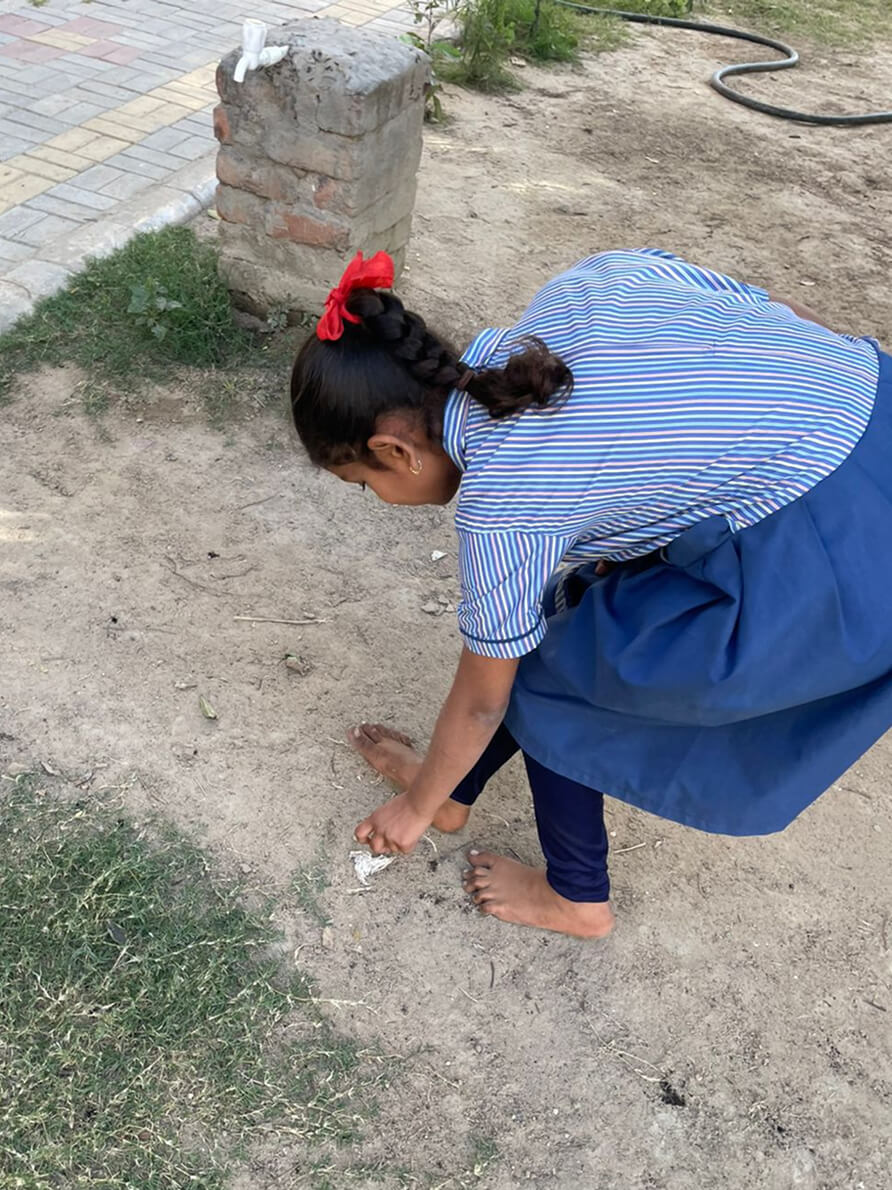 A schoolgirl in uniform crouching to pick up litter from the ground outdoors